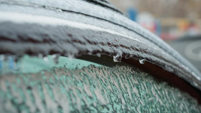 Close-up of frosted car window with melting icicles, blurred colorful background, highlighting texture contrasts and the vivid cold of winter scenery