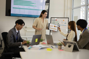 A group of people are working together in a conference room. They are all smiling and seem to be enjoying their time. There are several laptops and papers on the table, and a TV is mounted on the wall