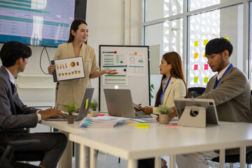 A group of people are working together in a conference room. They are all smiling and seem to be enjoying their time. There are several laptops and papers on the table, and a TV is mounted on the wall