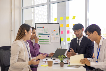 A group of people are working together in a conference room. They are all smiling and seem to be enjoying their time. There are several laptops and papers on the table, and a TV is mounted on the wall