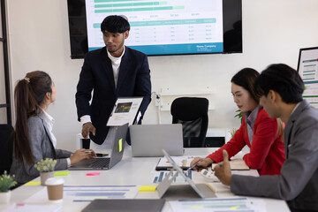A group of people are working together in a conference room. They are all smiling and seem to be enjoying their time. There are several laptops and papers on the table, and a TV is mounted on the wall