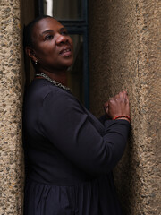 A confident woman stands between two textured walls, looking directly at the camera with a poised...