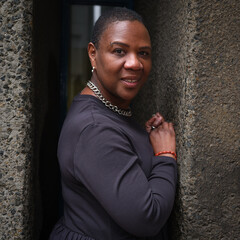 A confident woman stands between two textured walls, looking directly at the camera with a poised expression. She wears a dark dress, a silver chain necklace, and a red bracelet