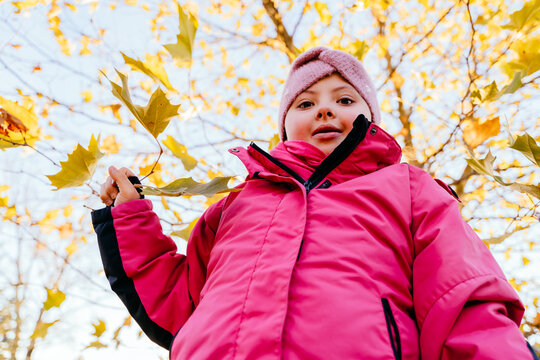 7-year-old girl with special needs in pink winter jacket holds yellow maple branch, looking up against bright autumn sky. Shot from low angle, highlighting energy and curiosity of childhood.