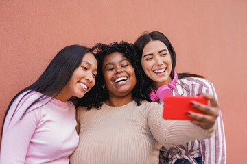 Young multiracial girls taking a selfie using mobile phone in the city with neutral wall background - Diverse latin women having fun together