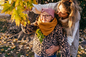 Girl with Down syndrome exploring autumn leaves mother in park, mother gently supporting her child,...