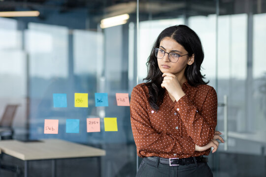 Serious thinking woman at workplace inside office standing near window. Businesswoman in glasses pondering and planning future startups and victories.