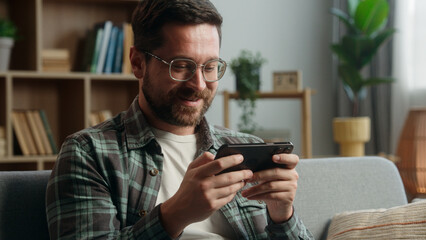 Caucasian man male guy user gamer at home video game addict browsing mobile phone play app playing smartphone shock emotional entertainment at home couch sofa apartment gaming online internet web fun © Yuliia