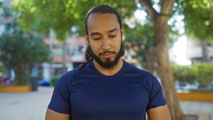 Young man with beard standing outdoors in an urban park with trees and buildings in the background,...
