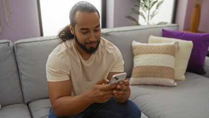 Obraz premium Young man with beard relaxing indoors holding smartphone in cozy apartment interior with modern decor casual style.