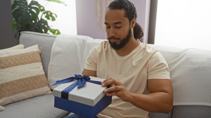 Fototapeta premium Young man with beard opening gift box in cozy living room setting, seated on sofa, creating a calm and thoughtful atmosphere.