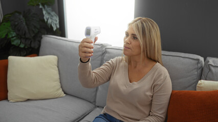 Woman sitting indoors using digital thermometer in modern living room, focusing intently on the...