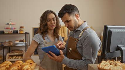 Man and woman wearing aprons working together in a bakery, interacting with a tablet while surrounded by freshly baked goods and a computer in the shop interior.