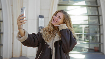 Fototapeta premium Woman smiling, taking selfie with smartphone on urban street, showcasing vibrant, youthful energy in city environment.