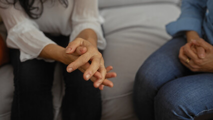 Women sitting together in a cozy living room, their hands clasped in a gesture of support and companionship, suggesting a moment of intimate conversation and connection.