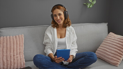 Obraz premium Young woman sitting on a couch in a living room wearing headphones and holding a tablet, smiling in a relaxed indoor setting with cushions and plants.
