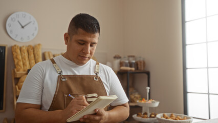 Young hispanic man in bakery writing notes in notebook indoors with pastries and breads visible in the background