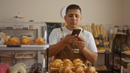 Young hispanic man using smartphone in bakery surrounded by pastries and breads on display shelves indoors