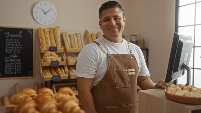 Young hispanic man in a bakery room smiling while wearing a brown apron and standing in front of shelves full of fresh bread and pastries