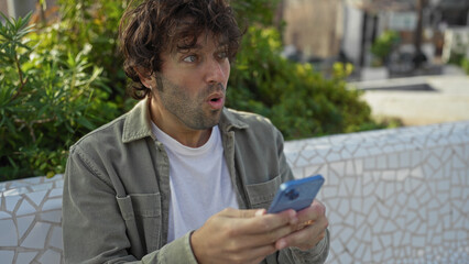 Fototapeta premium Young man with surprised expression holding smartphone casually sitting in urban park surrounded by greenery and cityscape background