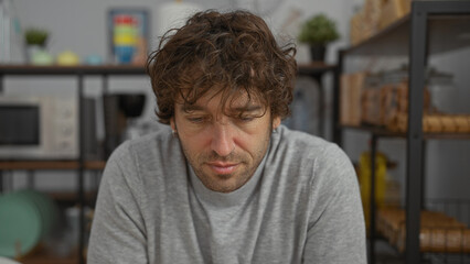 Young man with curly hair wearing gray sweater in an office setting with shelves behind him, looking down thoughtfully.