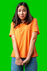 Young woman in orange shirt looks uncertain against bright green background in indoor setting