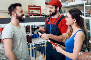 Salesman in red shirt and baseball cap is showing couple of clients new hammer in power tools store.
