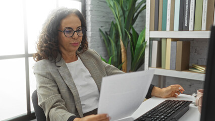 Middle-aged woman in office reading document at desk with keyboard, showcasing a mature professional setting with books and indoor plant decor.