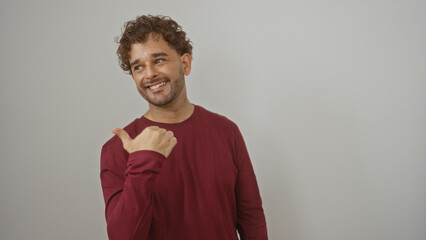 Young hispanic man smiling and pointing sideways with thumb over isolated white background