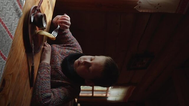 Vertical video of preteen boy eating fried potato pancakes with sour cream in cafe. Child dips national Slavic dish draniki into gravy boat and eats.