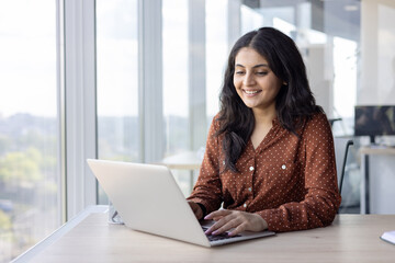 Young successful woman working inside office with laptop. Confident and happy businesswoman smiling typing on keyboard, sitting at desk near window.