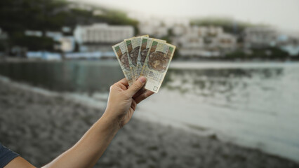 Man holding polish zloty banknotes by the seaside, showcasing currency against a beach and water backdrop, creating a sense of travel and finance outdoors.