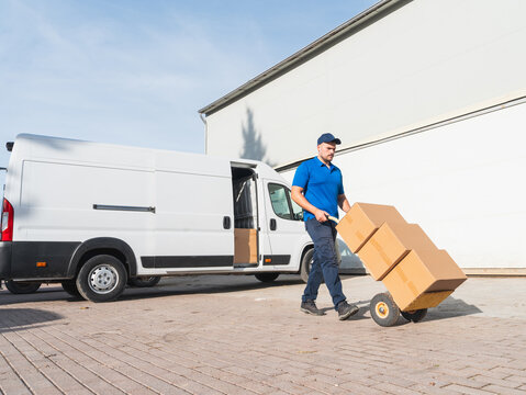 Delivery worker transporting boxes with a hand truck. Courier in a blue uniform moving cardboard boxes with a hand truck next to a delivery van. Logistics and shipping concept.