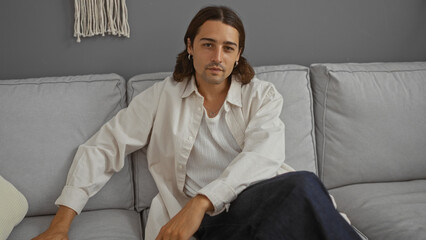 Fototapeta premium Young hispanic man sitting on a couch in a modern living room, exuding a calm and composed demeanor.