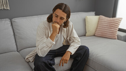 Fototapeta premium Handsome young hispanic man with long hair sitting thoughtfully on a couch in a modern living room