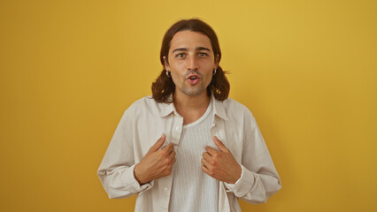 Young man with long hair points to himself in surprise against an isolated yellow background,...
