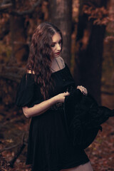 This striking photograph features a woman standing in a dark, moody forest, exuding a gothic and mysterious aura. She is dressed in an all-black ensemble, including a sheer, flowing cape.