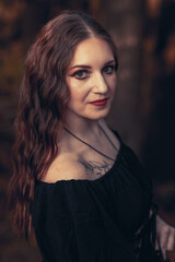 This striking photograph features a woman standing in a dark, moody forest, exuding a gothic and mysterious aura. She is dressed in an all-black ensemble, including a sheer, flowing cape.