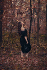 This striking photograph features a woman standing in a dark, moody forest, exuding a gothic and mysterious aura. She is dressed in an all-black ensemble, including a sheer, flowing cape.