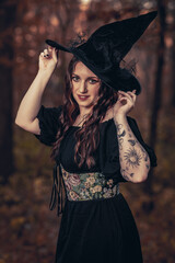 This striking photograph features a woman standing in a dark, moody forest, exuding a gothic and mysterious aura. She is dressed in an all-black ensemble, including a sheer, flowing cape.