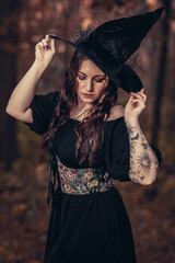 This striking photograph features a woman standing in a dark, moody forest, exuding a gothic and mysterious aura. She is dressed in an all-black ensemble, including a sheer, flowing cape.