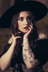 This striking photograph features a woman standing in a dark, moody forest, exuding a gothic and mysterious aura. She is dressed in an all-black ensemble, including a sheer, flowing cape.