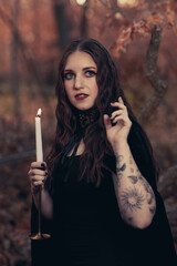 This striking photograph features a woman standing in a dark, moody forest, exuding a gothic and mysterious aura. She is dressed in an all-black ensemble, including a sheer, flowing cape.