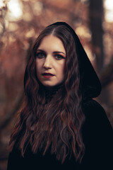 This striking photograph features a woman standing in a dark, moody forest, exuding a gothic and mysterious aura. She is dressed in an all-black ensemble, including a sheer, flowing cape.
