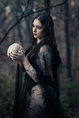 This striking photograph features a woman standing in a dark, moody forest, exuding a gothic and mysterious aura. She is dressed in an all-black ensemble, including a sheer, flowing cape.