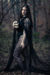 This striking photograph features a woman standing in a dark, moody forest, exuding a gothic and mysterious aura. She is dressed in an all-black ensemble, including a sheer, flowing cape.