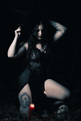 This striking photograph features a woman standing in a dark, moody forest, exuding a gothic and mysterious aura. She is dressed in an all-black ensemble, including a sheer, flowing cape.