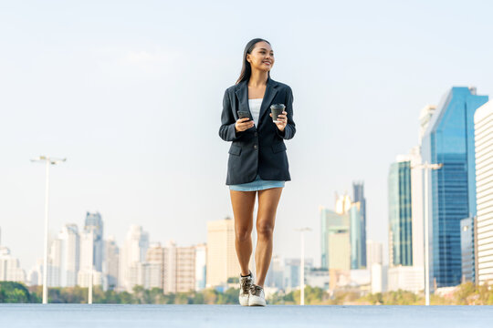 Young Asian Businesswoman Holding Phone and Coffee Walking in Modern City Outdoors