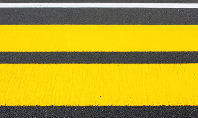 Fototapeta premium Close-Up View of Yellow Pedestrian Crosswalk Lines on a Black Asphalt Road with White Stripe in Urban Environment at Daytime