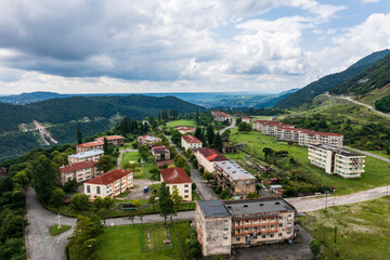 Obraz premium Aerial view of an abandoned residential settlement in the mountains. Rows of empty apartment buildings contrast with the surrounding green landscape, creating a post-apocalyptic atmosphere.